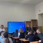 Классная комната: группа старшеклассников за партами слушает двух взрослых у большого экрана/монитора; видно часть учебной мебели и дверь.
