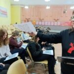 В зале школьники сидят в рядах с распечатками, на которых видны QR‑коды; ведущая протягивает лист с QR‑кодом.
