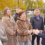 Мужчина беседует с группой жителей на вымощенной плиткой аллее сквера; на заднем плане лавочки, навесы и деревья с осенней листвой.