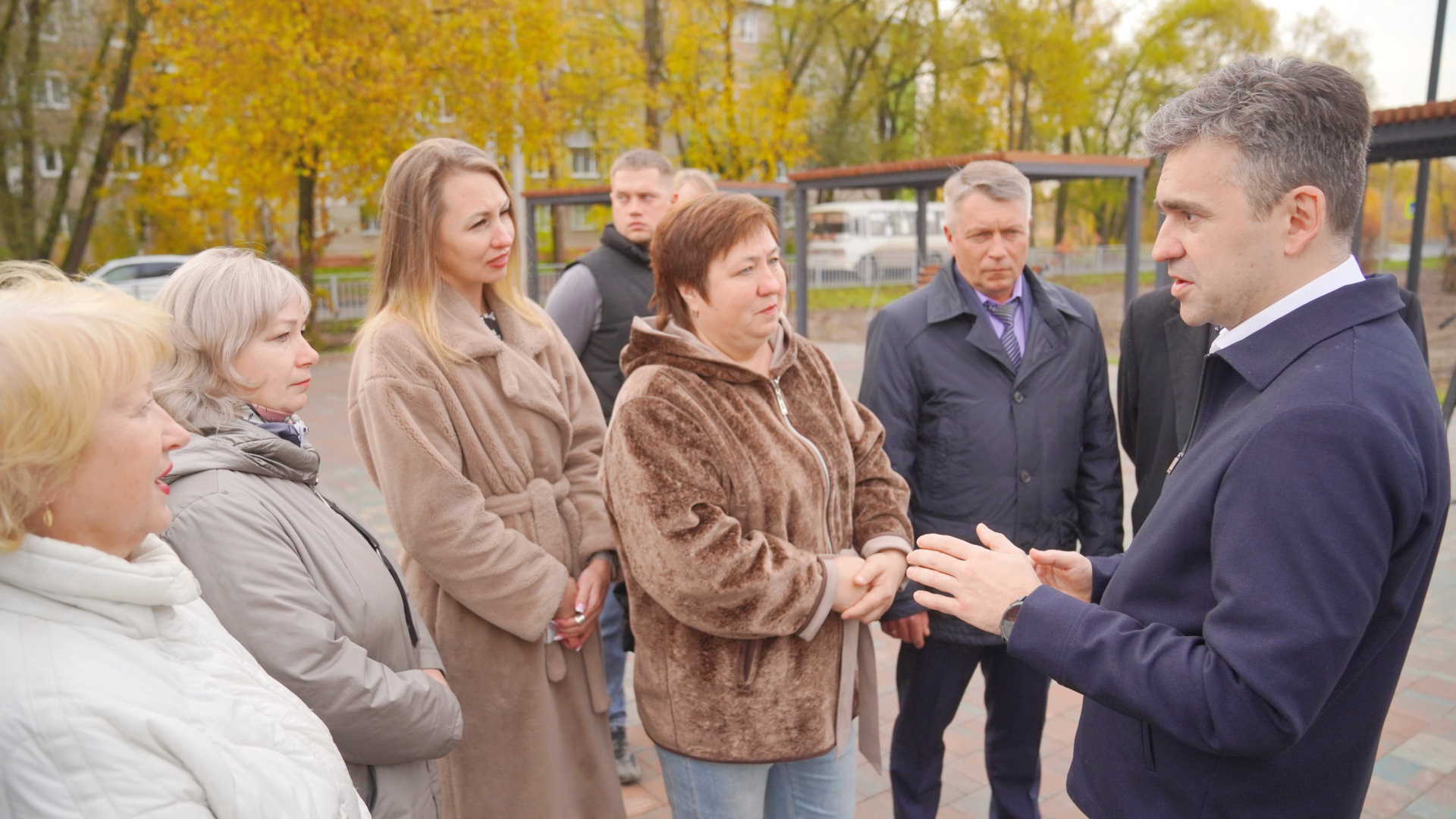 Мужчина беседует с группой жителей на вымощенной плиткой аллее сквера; на заднем плане лавочки, навесы и деревья с осенней листвой.