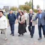 Группа людей идёт по вымощенной плиткой центральной аллее сквера; по краям видны лавочки, урны и фонарные столбы.