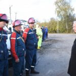 В ряд выстроены сотрудники экстренных и коммунальных служб в спецформе и защитных касках на открытой площадке; на заднем плане служебные машины, напротив них стоит пожилой мужчина в пальто.