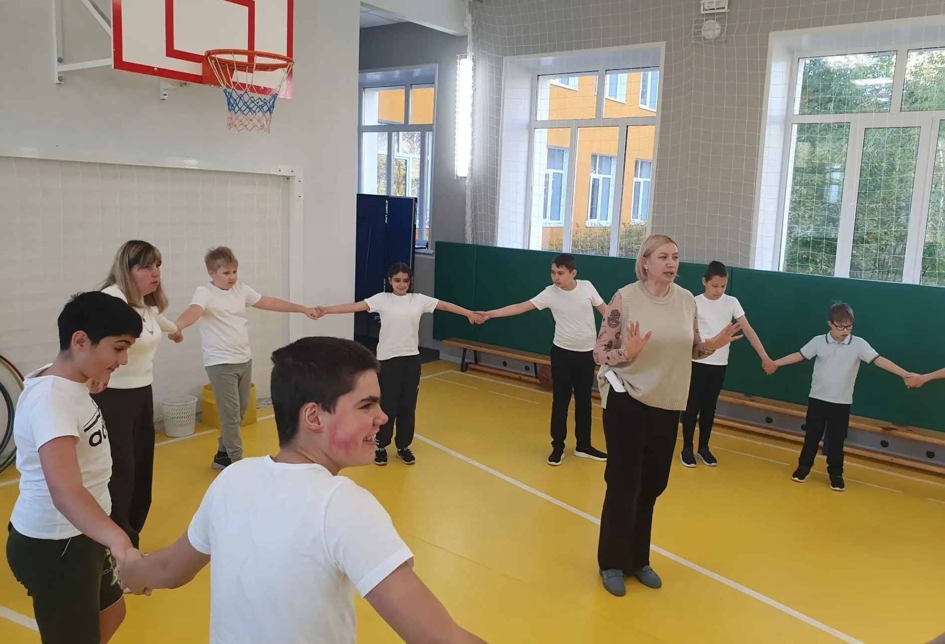 Дети и взрослые в спортивном зале держатся за руки в кругу; видны баскетбольное кольцо, защитные маты, жёлтый пол и сетка на окнах.
