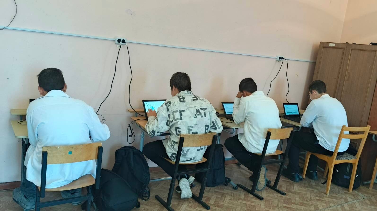 Несколько школьников-подростков сидят в классе за столами и проходят тесты на планшетах/ноутбуках.