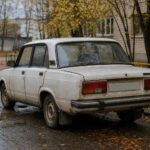 Старый грязный седан припаркован у жилого дома; вид сзади, осенняя дворовая улица