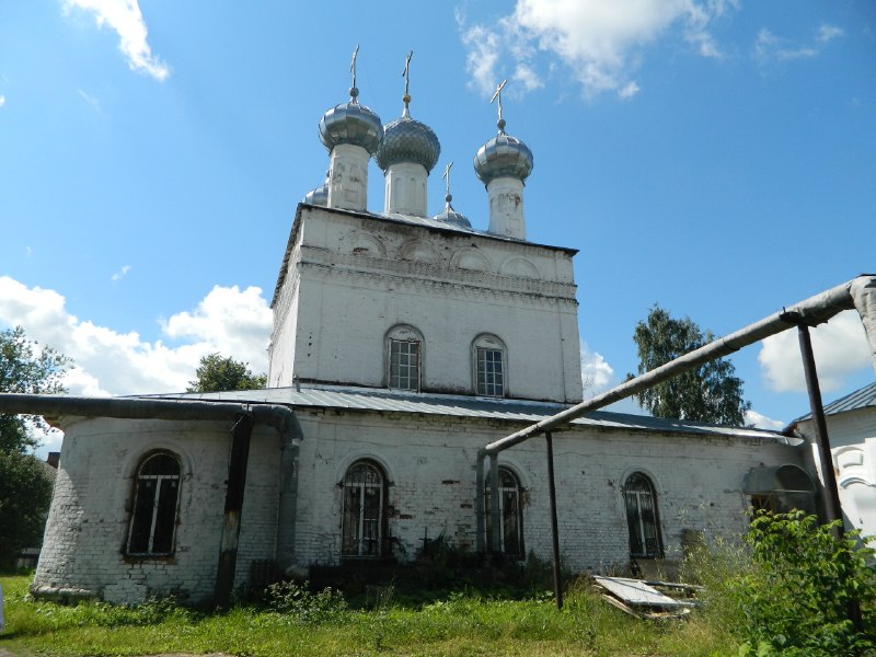 Белая кирпичная православная церковь с луковичными куполами (несколько глав), невысокой пристройкой спереди; летний пейзаж, трава и временные металлические опоры на переднем плане.