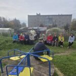 Группа жителей сидит и стоит на зелёной площадке с детской каруселью на переднем плане; на заднем плане — заборы, теплицы и жилые дома.