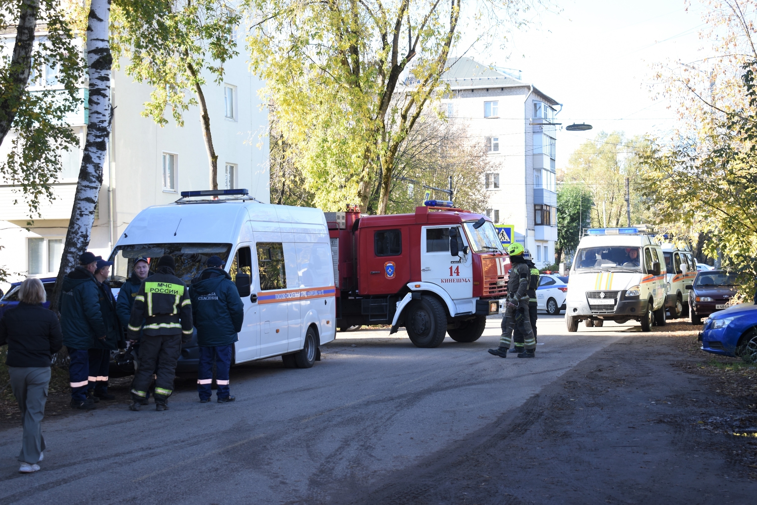 Улица с пожарным автомобилем (номер 14, надпись «КИНЕШМА»), аварийным фургоном и сотрудниками спасательных служб в форме с надписью «МЧС РОССИИ», рядом аварийные/медицинские автомобили.