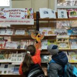Стеллаж с книжной выставкой: плакат «Самоцветная радуга России», баннер «Дни российской культуры», раздел «Глиняная сказка», книги о народных промыслах; внизу два ребёнка рассматривают экспозицию.