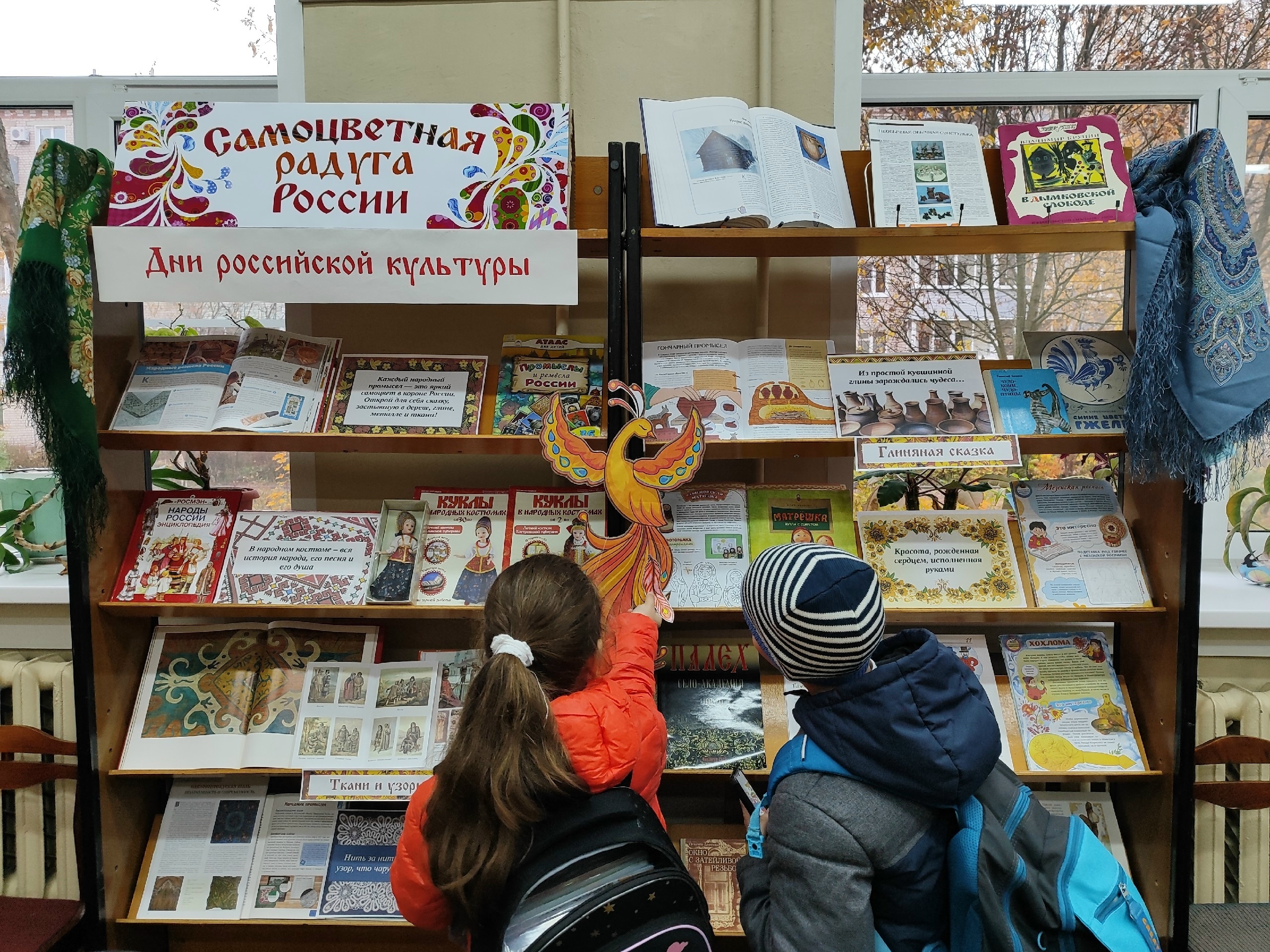 Стеллаж с книжной выставкой: плакат «Самоцветная радуга России», баннер «Дни российской культуры», раздел «Глиняная сказка», книги о народных промыслах; внизу два ребёнка рассматривают экспозицию.
