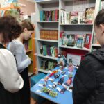 Посетители осматривают выставочный стол с тряпичными куклами в библиотеке; на заднем плане книжные стеллажи и вывеска с названием выставки.