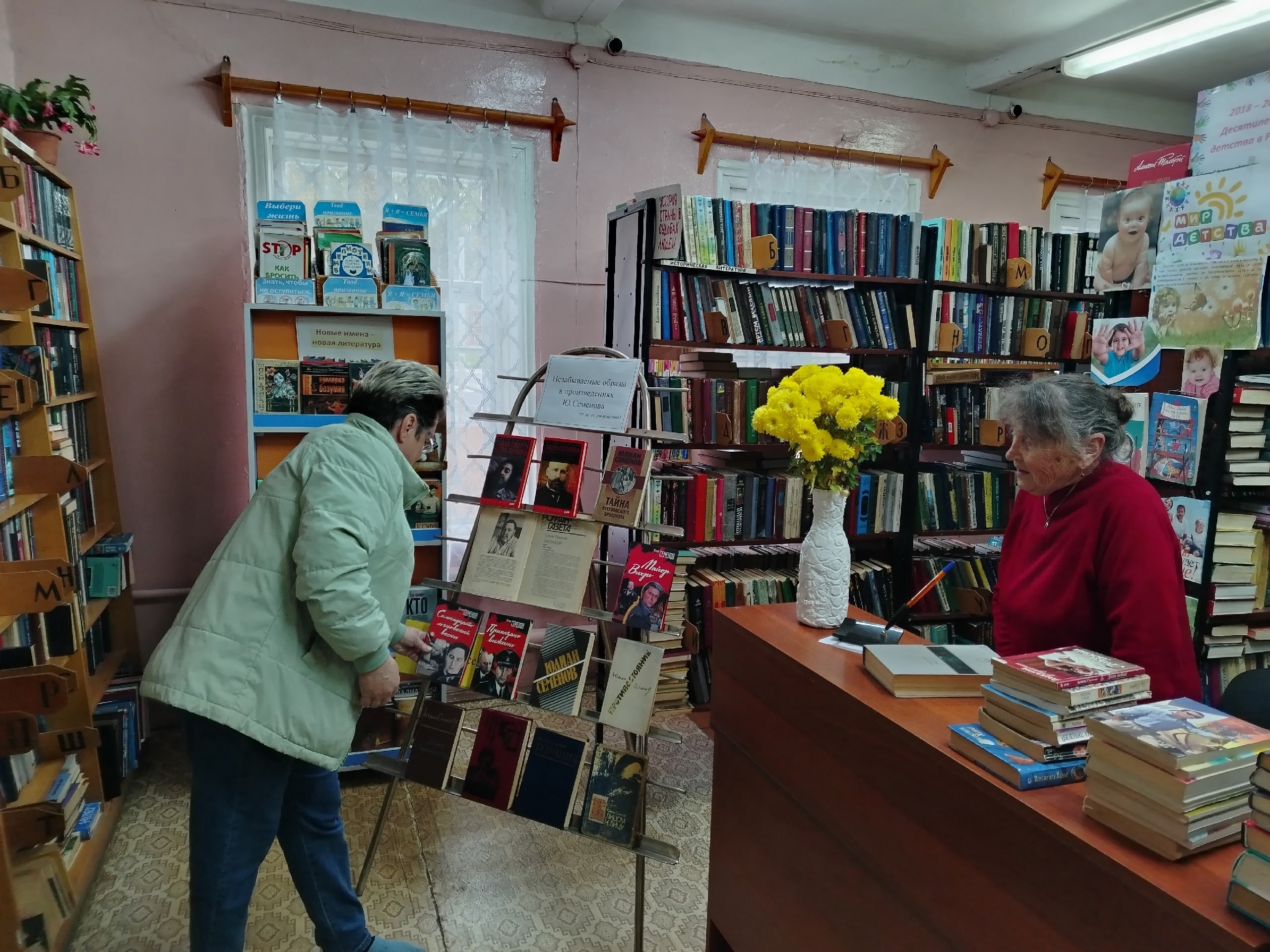 Книжная выставка в библиотеке: стенд с книгами и вывеской об экспозиции Ю. Семёнова, книжные полки и посетители