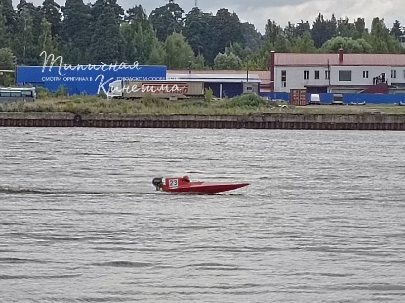 Красная гоночная моторная лодка с номером 23 плывёт по реке; на заднем плане берег с набережной/промышленными постройками и деревьями.