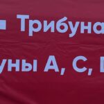 Красный баннер/плакат с белыми стрелками и указателями трибун: надпись «Трибуна В» и фрагмент «рибуны A, C, D» (навигация по трибунам стадиона).