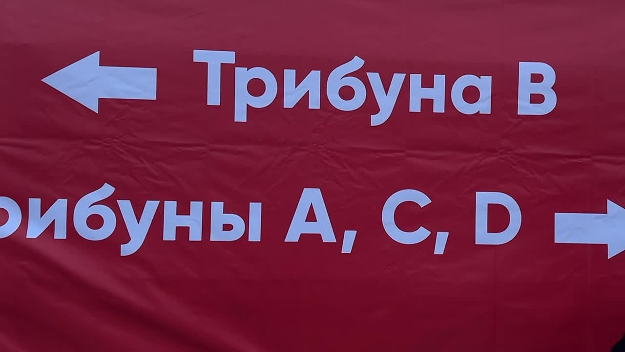 Красный баннер/плакат с белыми стрелками и указателями трибун: надпись «Трибуна В» и фрагмент «рибуны A, C, D» (навигация по трибунам стадиона).