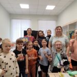 Группа детей и взрослых в зале мастерской: дети держат кисти, на столах видны рисунки, в витрине — выставленные работы.