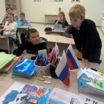 Младшие школьники в классе за партами во время занятия; на переднем плане учебники и распечатки, одна из которых — иллюстрация с ребёнком и мобильным телефоном/монстром.