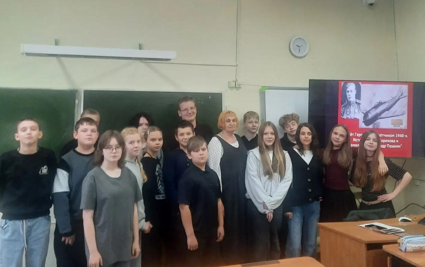 Группа школьников с учителем в классе на музейном уроке; на экране справа виден портрет лётчика и изображение самолёта