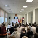 Школьное собрание в актовом/малом зале: ряд учащихся, сидящих лицом к ведущей у экрана; на стене герб, рядом флаги.