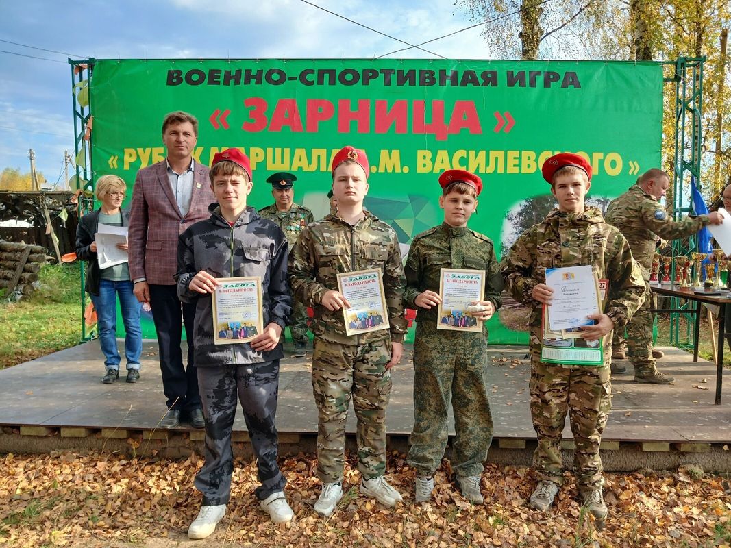 Юнармейцы-подростки в камуфляжной форме и красных беретах с грамотами стоят на сцене перед зелёным баннером с надписью «Зарница» и названием рубежа Василевского.