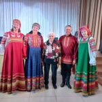 Группа солистов в народных костюмах и пожилая женщина на сцене
